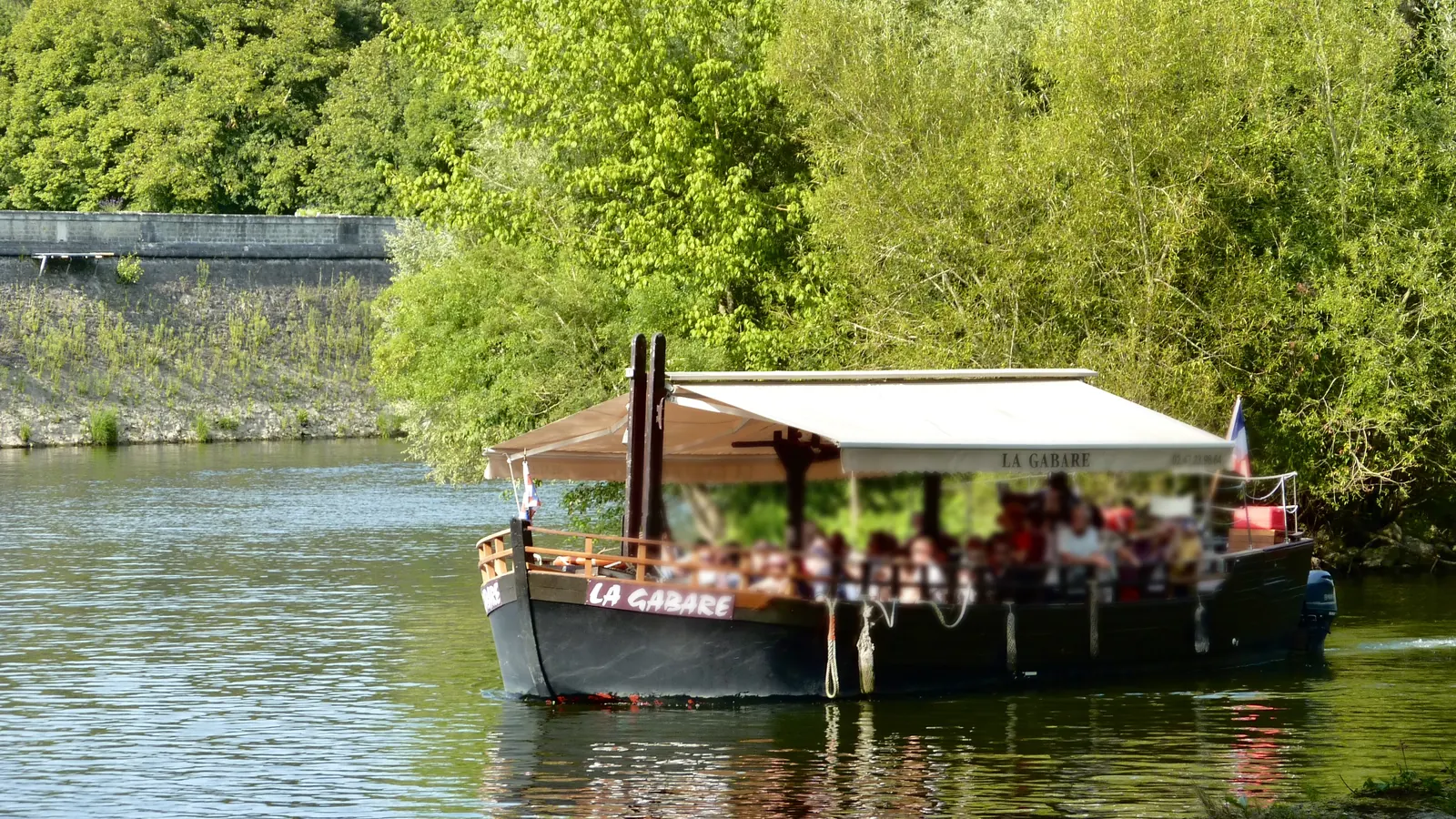La gabare de promenade sur le Cher, sa bâche claire tendue au-dessus du pont, le long d’une berge boisée.